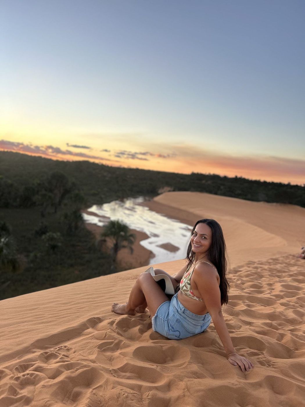 Isadora sentada nas Dunas do Jalapão ao pôr do sol, com o rio ao fundo